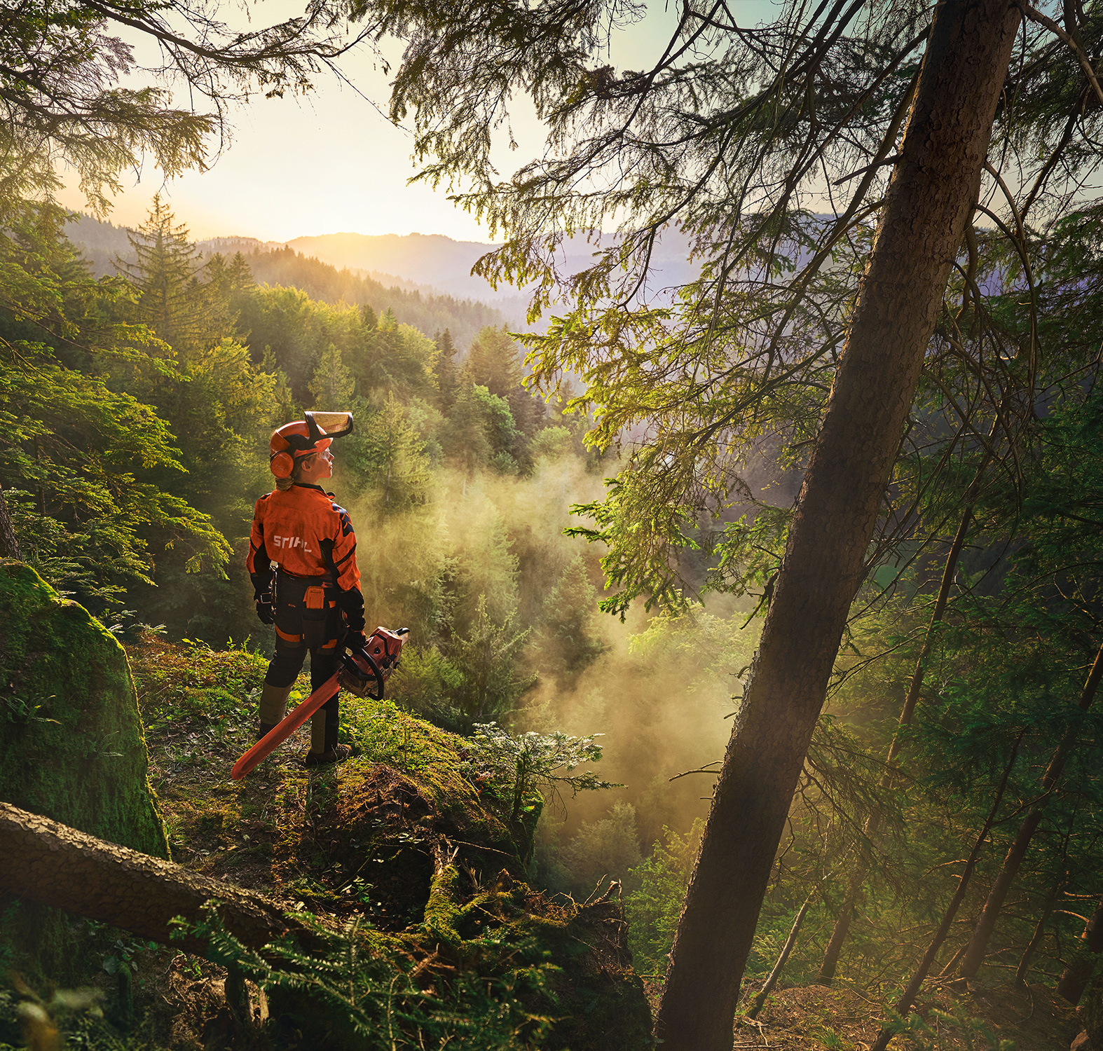 Una donna è in piedi sul bordo di una scogliera in una foresta di un verde saturo. Indossa l'abbigliamento da lavoro arancione STIHL, la cuffia ADVANCE ProCom e tiene in mano una motosega STIHL MS. 