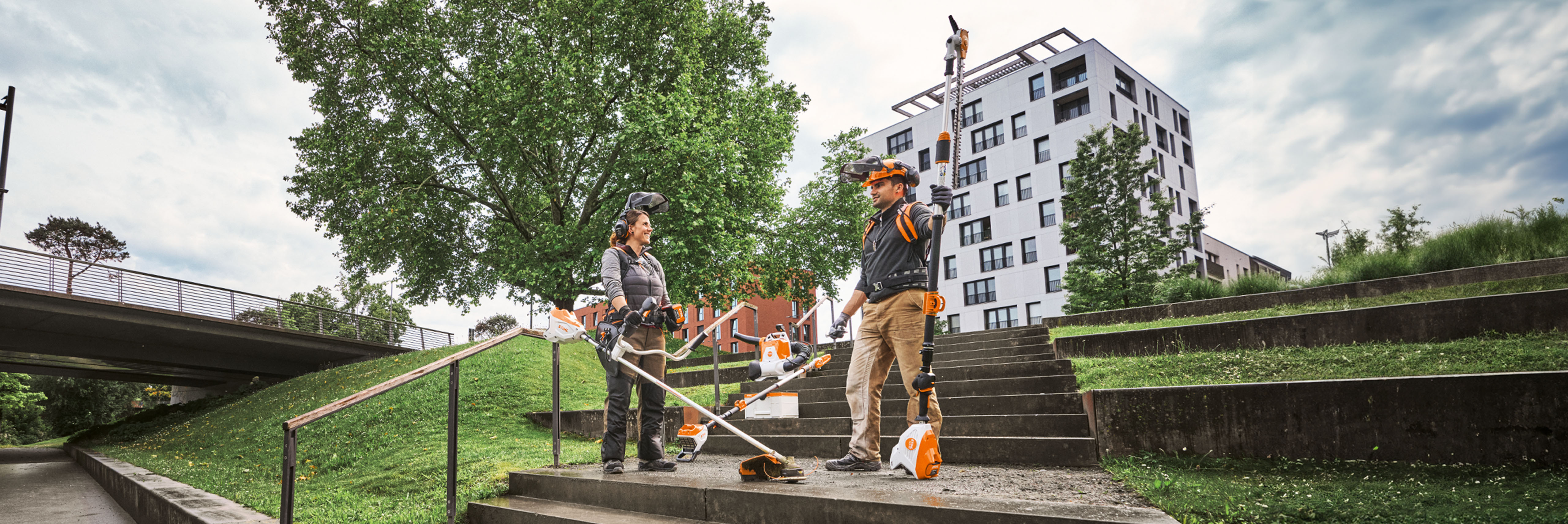 Due professionisti della paesaggistica che utilizzano gli attrezzi e le batterie STIHL Pro su un'area verde urbana con scale e alberi.