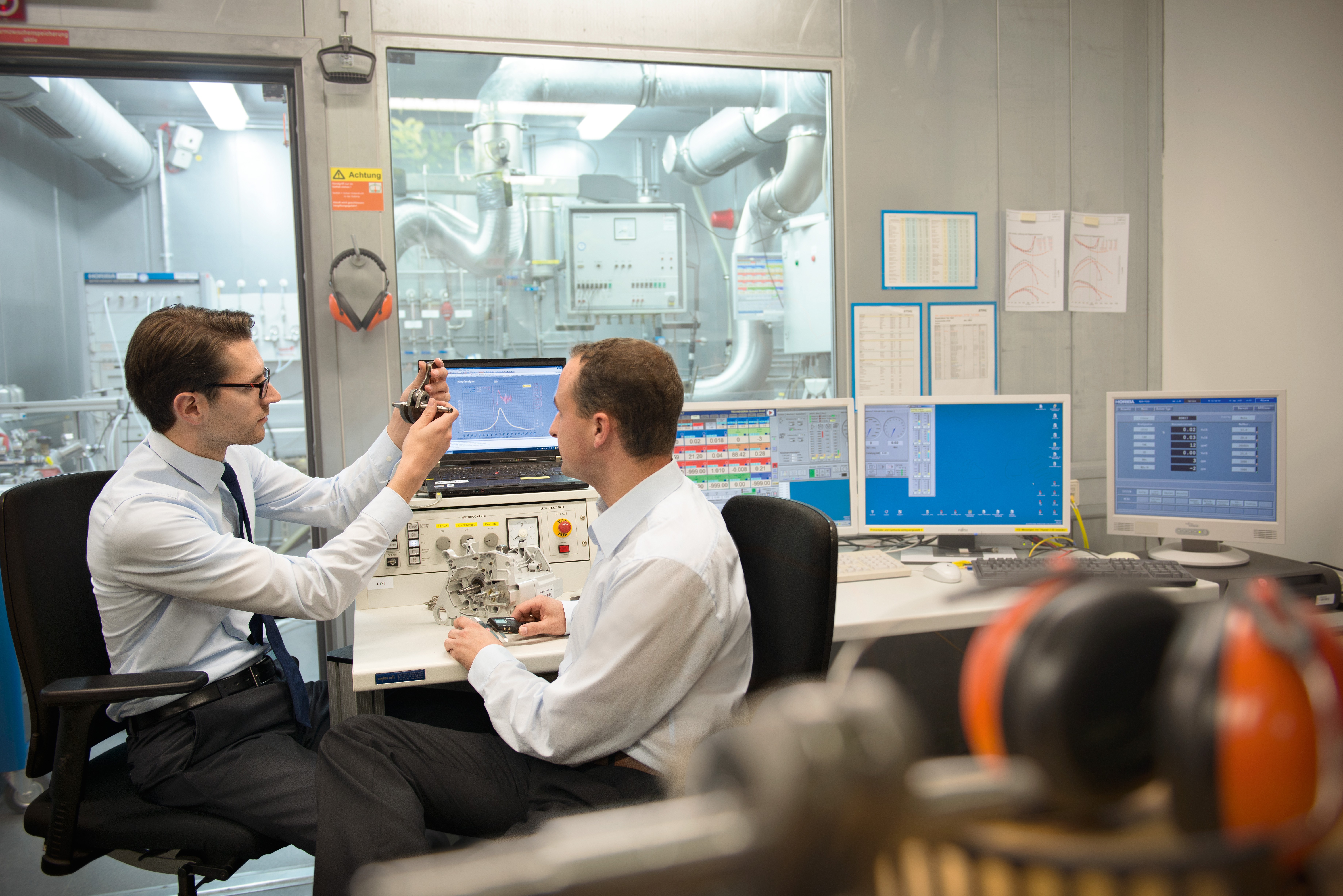Due uomini siedono in un laboratorio a una scrivania con diversi monitor ed esaminano un componente di un nuovo prodotto