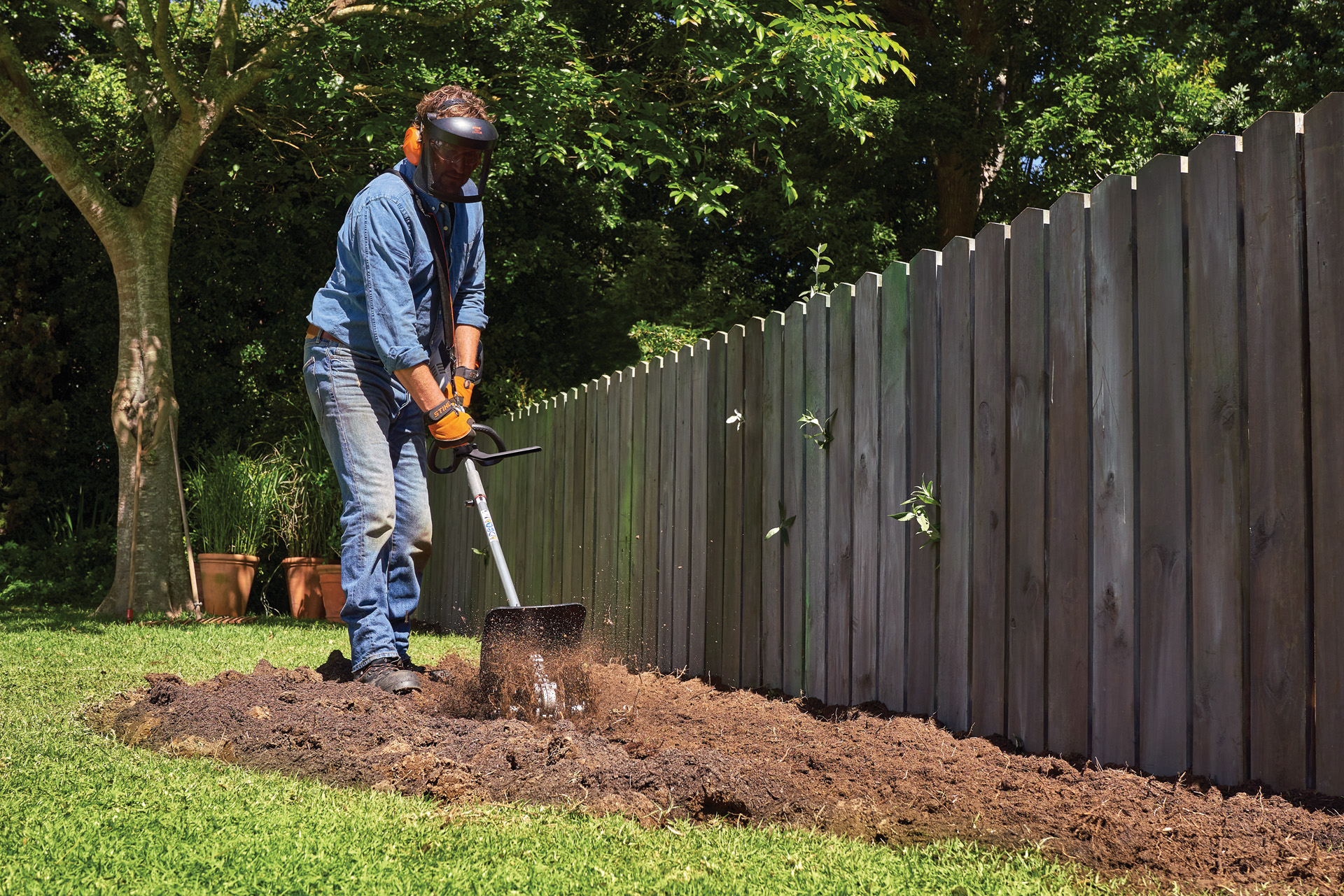 Persona munita di visiera e protezioni acustiche che scava una superficie in giardino utilizzando lo STIHL KombiSystem con fresa per terreno allo scopo di realizzare un'aiuola