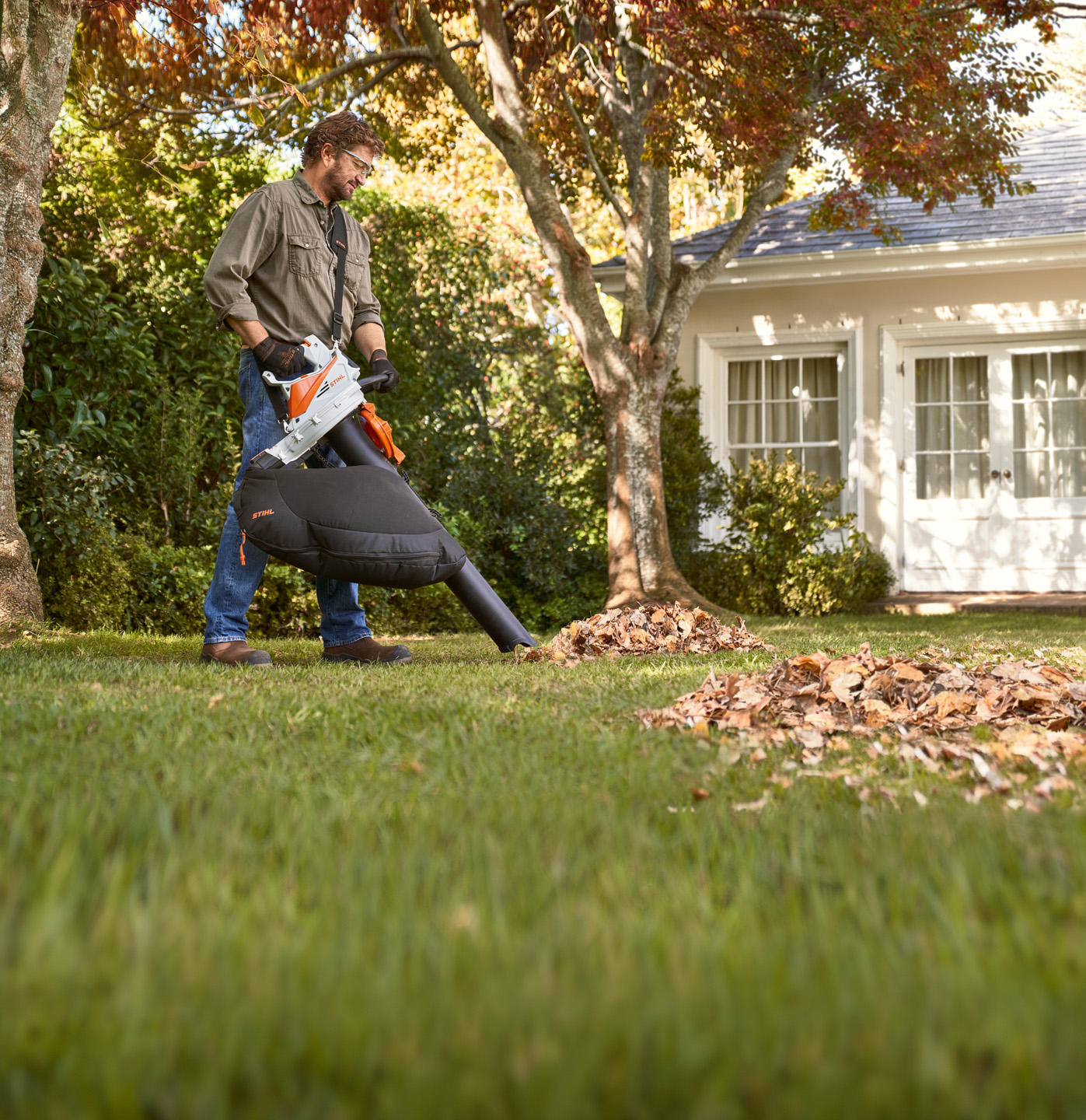 Uomo che aspira le foglie davanti a un albero in giardino con l'aspiratore-trituratore a batteria STIHL SHA 56