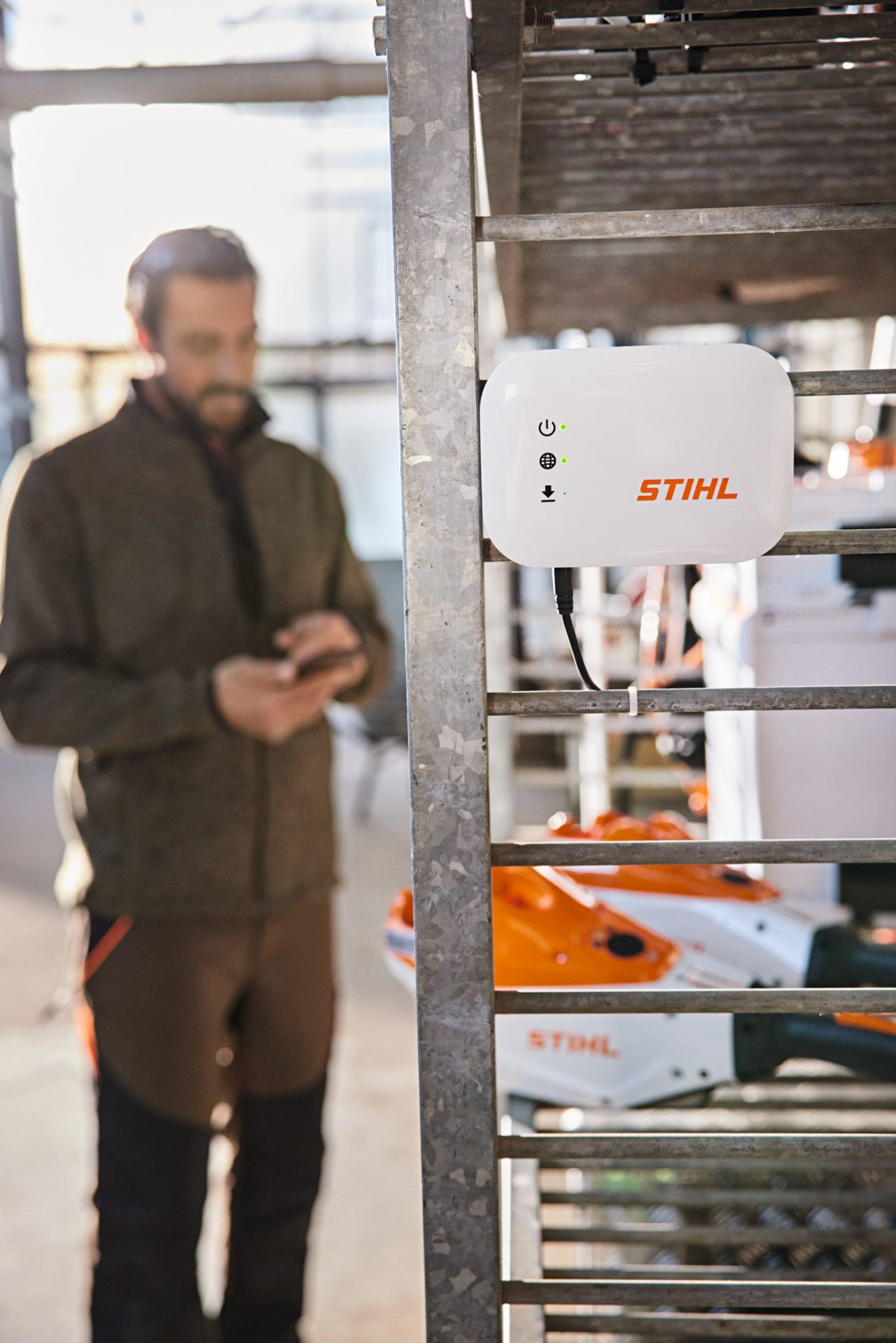Una STIHL connected Box è appesa a uno scaffale dell'officina. Sullo sfondo si vede un giardiniere e paesaggista che interagisce con il proprio smartphone. 