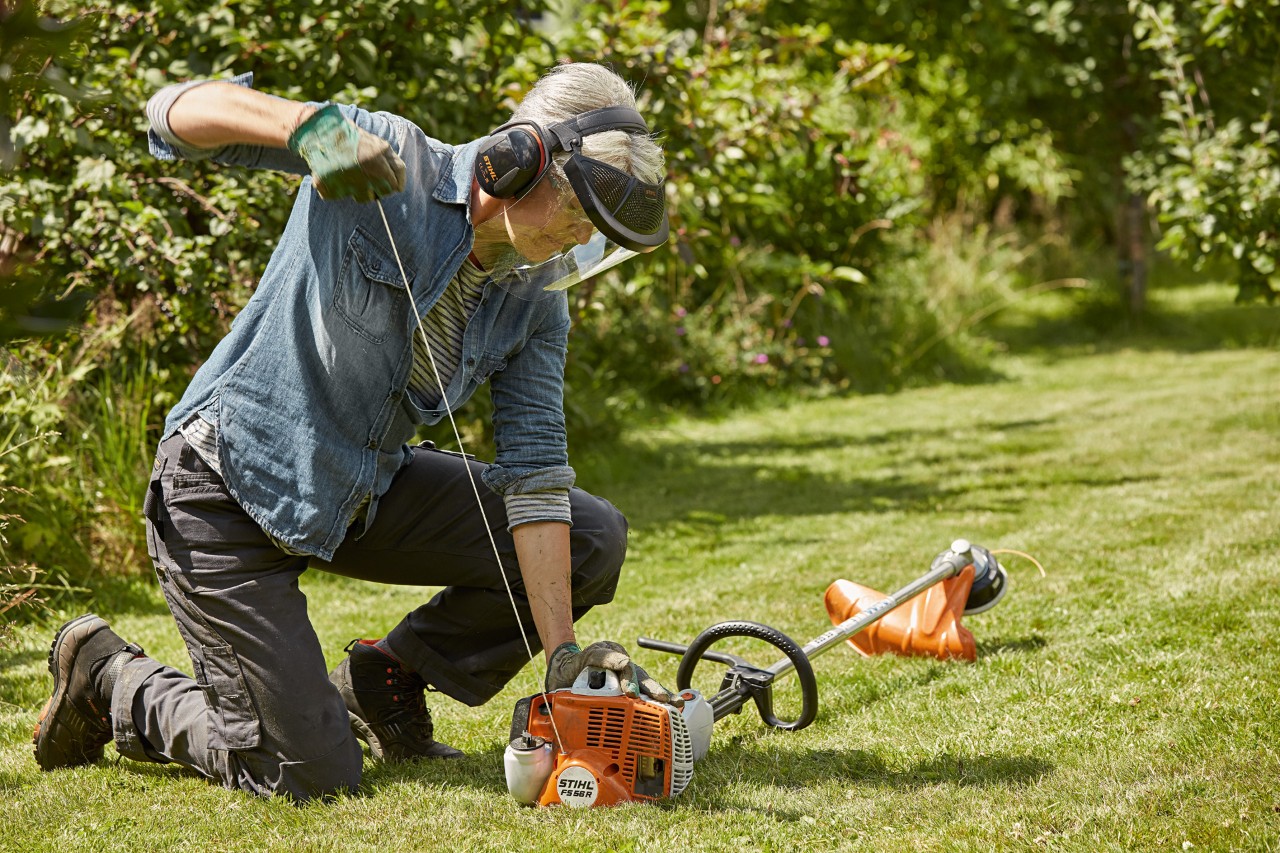 Donna con protezione auricolare e schermo per il viso che sta per avviare un decespugliatore a benzina STIHL FS 56 R su un terreno verde